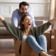 Happy family celebrating moving day, playful young couple having fun at home, funny activity, laughing husband pushing excited wife sitting in cardboard box