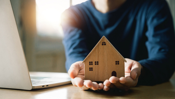 Hands holding a wooden model home with an open laptop Hands holding a wooden model home with an open laptop