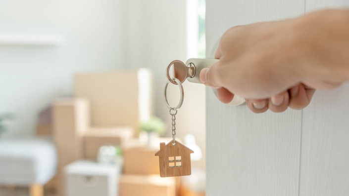 A key inserted into a door of the new house with a hand opening it, inside the room are cardboard boxes and furniture A key inserted into a door of the new house with a hand opening it, inside the room are cardboard boxes and furniture